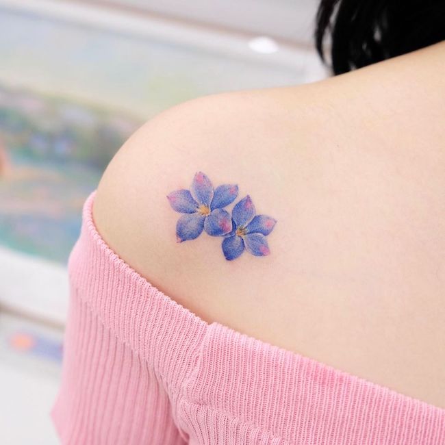 Piccolo fiore blu e viola tatuato sul retro della spalla da @siyeon_tattoo